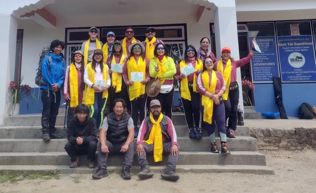 Black Yak Expedition team group photo at Yuksom base, featuring tour proprietor, trekking guide, and trekkers wearing yellow ceremonial scarves after completing a Goechala