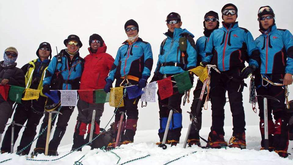 Black Yak Expeditions team led by founder Puspa Lal Gurung standing with international trekkers on a Himalayan summit, equipped with ice axes and climbing gear, with prayer flags and snow-covered high-altitude landscape in the background,